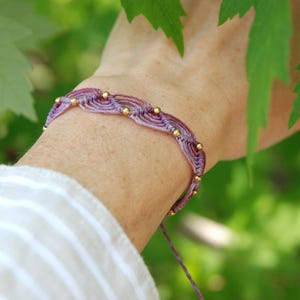May include: A handmade macrame bracelet in shades of purple and mauve, adorned with small gold beads. The bracelet is displayed on a wrist, with a blurred green foliage background. The bracelet is secured with a sliding knot closure.
