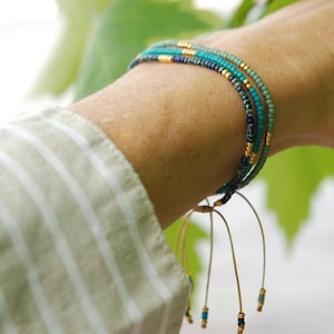 以下が含まれることがあります： ターコイズ、ネイビー、ゴールドの色合いのマルチストランドビーズブレスレットが手首に着用されています。 ブレスレットは小さなビーズでできており、繊細で層状の外観をしています。 背景には緑の葉とストライプのシャツがあります。