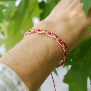 May include: A handmade macrame bracelet in shades of pink and red, accented with small gold-colored beads. The bracelet is worn on a wrist, with green leaves in the background. The bracelet is a delicate piece of jewelry.