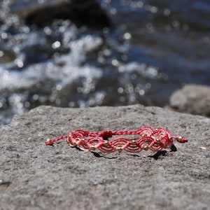 Pu&ograve; includere: Braccialetto in macram&egrave; fatto a mano, rosso e crema, ornato da piccole perline dorate. Appoggiato su una roccia grigia, con acqua sfocata sullo sfondo. Un gioiello artigianale.