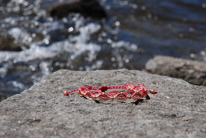 Pu&ograve; includere: Braccialetto in macram&egrave; fatto a mano, rosso e crema, ornato da piccole perline dorate. Appoggiato su una roccia grigia, con acqua sfocata sullo sfondo. Un gioiello artigianale.