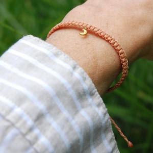May include: A peach-coloured braided cord bracelet featuring a small, gold-toned crescent moon charm. The bracelet is constructed with a series of intricate knots and includes adjustable cord ends that hang loosely from the main band.