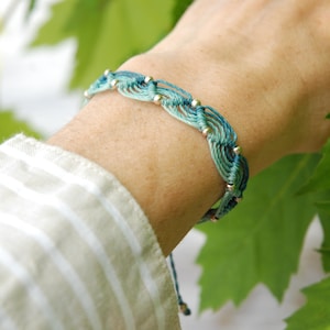 May include: A teal and blue macrame bracelet with gold beads. The woven design is visible, and the bracelet is tied with a matching cord. The bracelet is shown on a wrist. The bracelet is a piece of handmade jewellery.