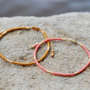 Könnte beinhalten: Zwei verstellbare Armbänder aus kleinen runden Perlen. Ein Armband besteht aus senfgelben Perlen mit eingearbeiteten goldfarbenen Metallakzenten, während das andere korallenrosa Perlen mit ähnlichen goldfarbenen Metallakzenten aufweist.