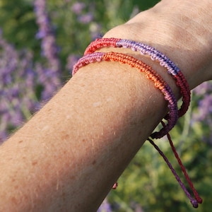 May include: Two handmade macrame bracelets in shades of red, orange, and purple are worn on a wrist. The bracelets are knotted with a sliding knot closure. The background is a blurred view of purple flowers.
