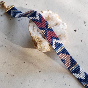 May include: A beaded bracelet with a geometric pattern in shades of blue, white, red, and gold. The bracelet is laid across a white and tan stone, and has a gold clasp.