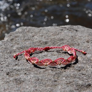 Pu&ograve; includere: Braccialetto in macram&egrave; fatto a mano, nei toni del rosso e crema, ornato da piccole perline dorate. Il braccialetto &egrave; appoggiato su una pietra grigia, con uno sfondo d'acqua sfocata.