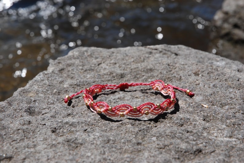 Pu&ograve; includere: Braccialetto in macram&egrave; fatto a mano, nei toni del rosso e crema, ornato da piccole perline dorate. Il braccialetto &egrave; appoggiato su una pietra grigia, con uno sfondo d'acqua sfocata.