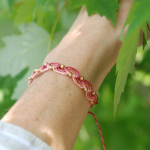 Pu&ograve; includere: Braccialetto in macram&egrave; rosa e oro. Il braccialetto presenta un design intrecciato con piccole perline dorate. Lo sfondo &egrave; verde tenue, con foglie nell'angolo in alto a destra. Un gioiello delicato.