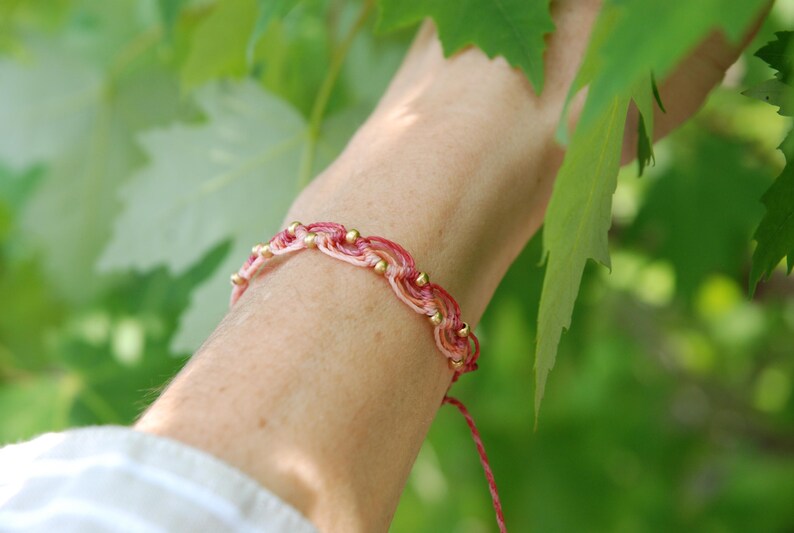 Pu&ograve; includere: Braccialetto in macram&egrave; rosa e oro. Il braccialetto presenta un design intrecciato con piccole perline dorate. Lo sfondo &egrave; verde tenue, con foglie nell'angolo in alto a destra. Un gioiello delicato.