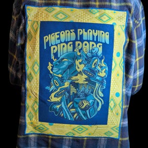 May include: Blue and yellow plaid flannel shirt with a large, rectangular patch on the back. The patch features a blue and yellow graphic design with the text "Pigeons Playing Ping Pong". The shirt has a classic button-down design.