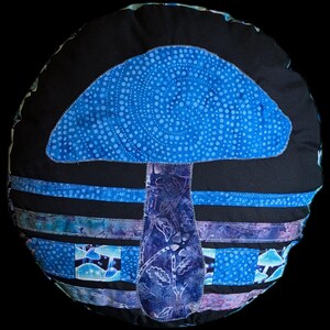 May include: Round, black pillow with a blue mushroom design. The mushroom cap is a lighter blue with white dots, and the stem is a darker blue with a leaf pattern. Blue and purple stripes accent the base.