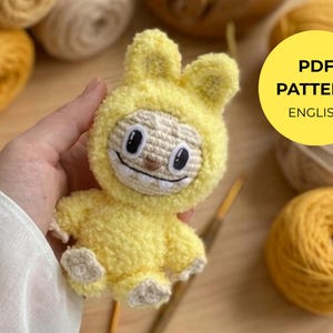May include: A handmade yellow crochet bunny toy with a smiling face and floppy ears. The toy is held in a person's hand, with yarn and crochet hooks in the background. A yellow circle with the text "PDF PATTERN ENGLISH" is in the image.