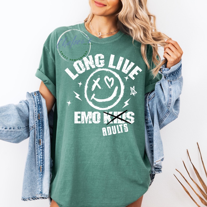 May include: Green t-shirt with the text "LONG LIVE EMO KIDS" in white. The design includes a smiley face with hearts for eyes and a crossed-out "S" in "KIDS", with lightning bolts and stars. The shirt has rolled-up sleeves.