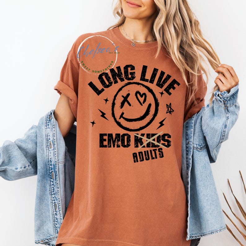 May include: A rust-colored t-shirt with the text "LONG LIVE EMO KIDS ADULTS" in black. The design includes a smiley face with crossed-out eyes and a heart, along with stars and lightning bolts. The shirt has rolled-up sleeves.