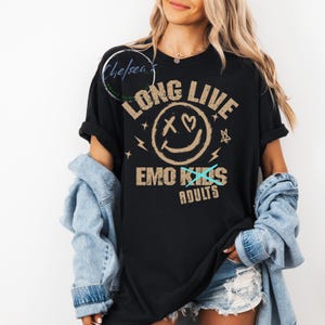 May include: Black t-shirt with the text "LONG LIVE EMO ADULTS" in a distressed, tan font. The design includes a smiley face with an X for eyes and a heart for a mouth. The shirt is worn with a denim jacket and shorts.