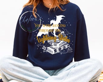 Op de afbeelding: Marineblauw sweatshirt met een witte afbeelding van een draak, boeken en de tekst "You Were Dancing Through The Lightning Strikes" in goud en wit. Een top met lange mouwen.