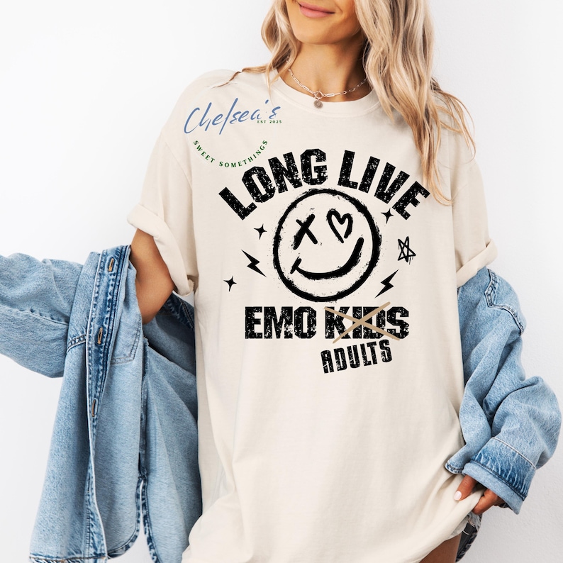 May include: Cream-colored t-shirt with the text "LONG LIVE EMO KIDS ADULTS" in black. The design includes a smiley face with an X for an eye and a heart, plus lightning bolts and stars.