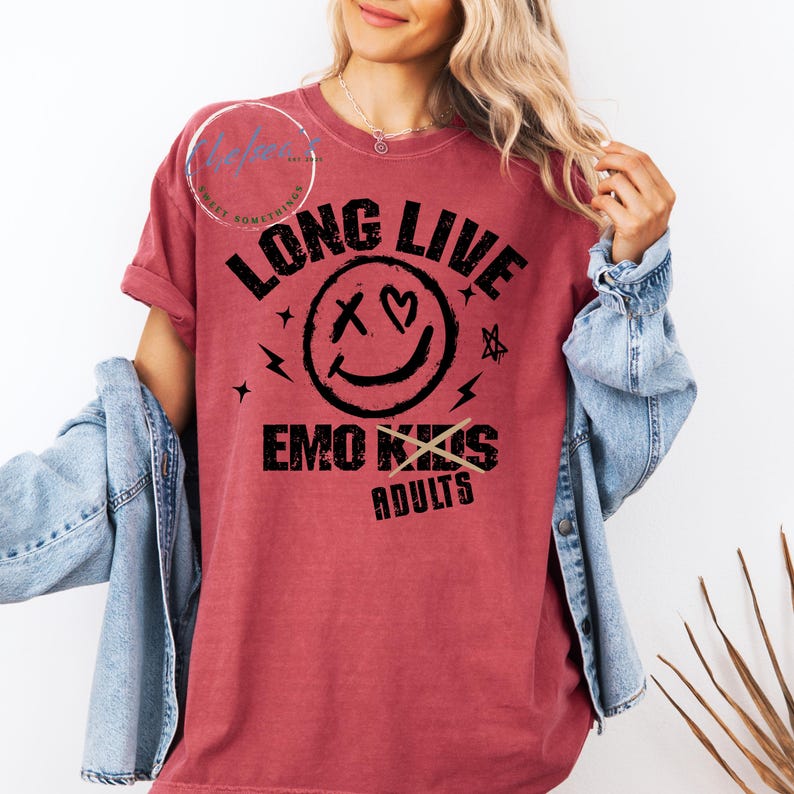 May include: A maroon t-shirt with the text "LONG LIVE EMO KIDS" above "ADULTS" with a smiley face graphic. The graphic includes a heart and an X for eyes. The shirt has rolled-up sleeves and is paired with a denim jacket.