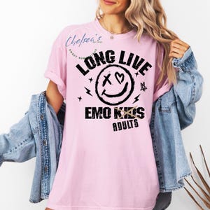 May include: A light pink t-shirt with the text "LONG LIVE EMO KIDS ADULTS" in black. The design includes a smiley face with an X for eyes and a heart, plus lightning bolts and stars. The shirt is a casual style.