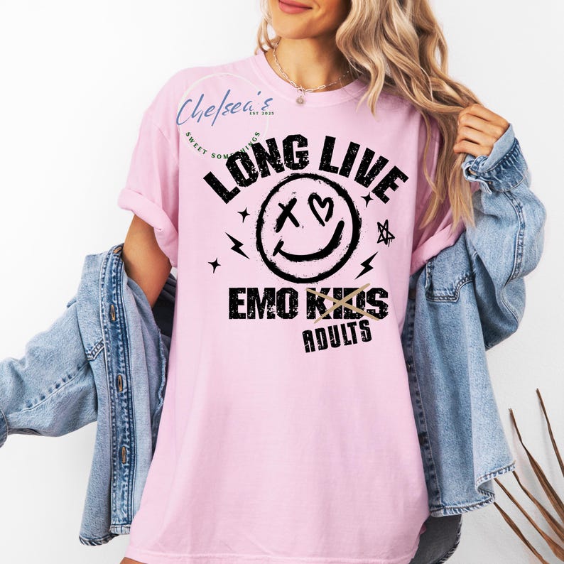 May include: A light pink t-shirt with the text "LONG LIVE EMO KIDS ADULTS" in black. The design includes a smiley face with an X for eyes and a heart, plus lightning bolts and stars. The shirt is a casual style.