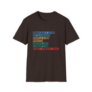 May include: A dark brown t-shirt with a white, blue, orange, and red graphic print that reads "Democracy Over Dictatorship, People Over Profits, Justice, Not Corruption, Resist. Persist. Rise."