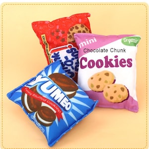 Pode incluir: Uma coleção de sacos de pelúcia para lanches de biscoitos em várias cores. Uma bolsa é vermelha com um gráfico de biscoito e texto. Outra é azul com o texto "YUMEO" e "Funny Stuffed Cookies!". Uma terceira é rosa com o texto "Chocolate Chunk Cookies" e ilustrações de biscoitos.