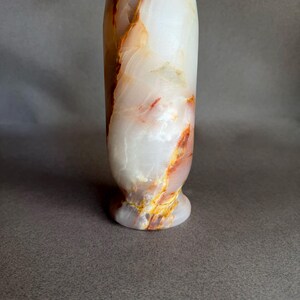 Pode incluir: Um vaso alto de &ocirc;nix branco com veios laranja e amarelos. O vaso tem uma base arredondada e um corpo cil&iacute;ndrico, ideal para exibir flores ou como pe&ccedil;a decorativa. A pedra natural tem um acabamento polido.