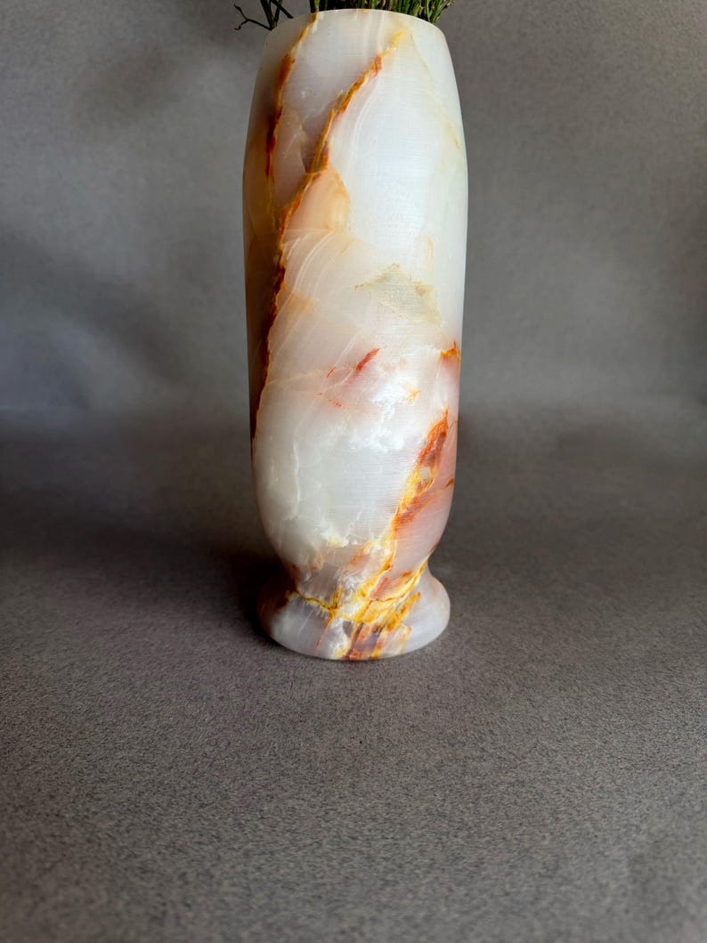 Pode incluir: Um vaso alto de &ocirc;nix branco com veios laranja e amarelos. O vaso tem uma base arredondada e um corpo cil&iacute;ndrico, ideal para exibir flores ou como pe&ccedil;a decorativa. A pedra natural tem um acabamento polido.