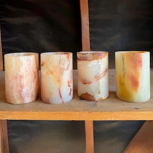 May include: Four cylindrical candle holders made of translucent stone. The holders display a range of colors, including cream, peach, and brown, with unique veining patterns. They are arranged on a wooden shelf.