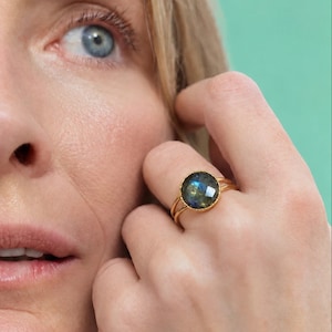 May include: A gold ring featuring a round, faceted labradorite gemstone with blue and grey iridescence. The stone is set in a simple gold bezel on a thin, polished gold band that splits into two delicate strands at the sides.