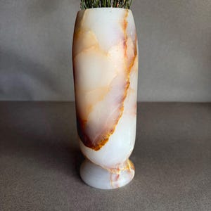 Pode incluir: Um vaso cil&iacute;ndrico de pedra branca e laranja-castanho. O vaso tem uma base arredondada e cont&eacute;m flores secas. A pedra tem um acabamento polido e o vaso tem cerca de 20 cm de altura.