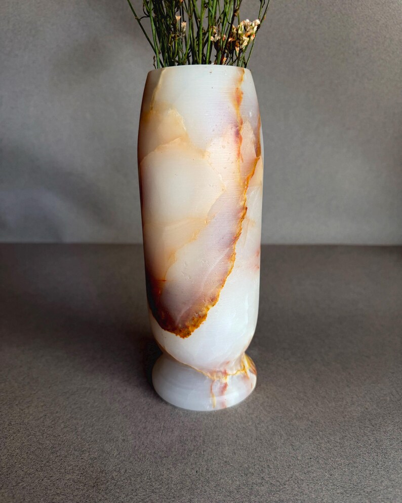 Pode incluir: Um vaso cil&iacute;ndrico de pedra branca e laranja-castanho. O vaso tem uma base arredondada e cont&eacute;m flores secas. A pedra tem um acabamento polido e o vaso tem cerca de 20 cm de altura.