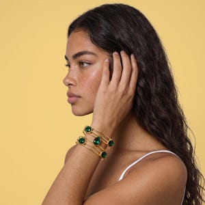 May include: A gold-toned cuff bracelet featuring three parallel bands and four faceted emerald green stones. The stones are arranged in a mix of round and teardrop shapes, set securely within the metal frame of the open-style wrist accessory.