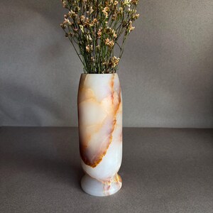 Pode incluir: Um vaso decorativo de &ocirc;nix branco e laranja com a parte superior alargada e uma base arredondada. O vaso &eacute; preenchido com flores secas com pequenas flores de cor clara. O vaso &eacute; um elemento decorativo para casa.