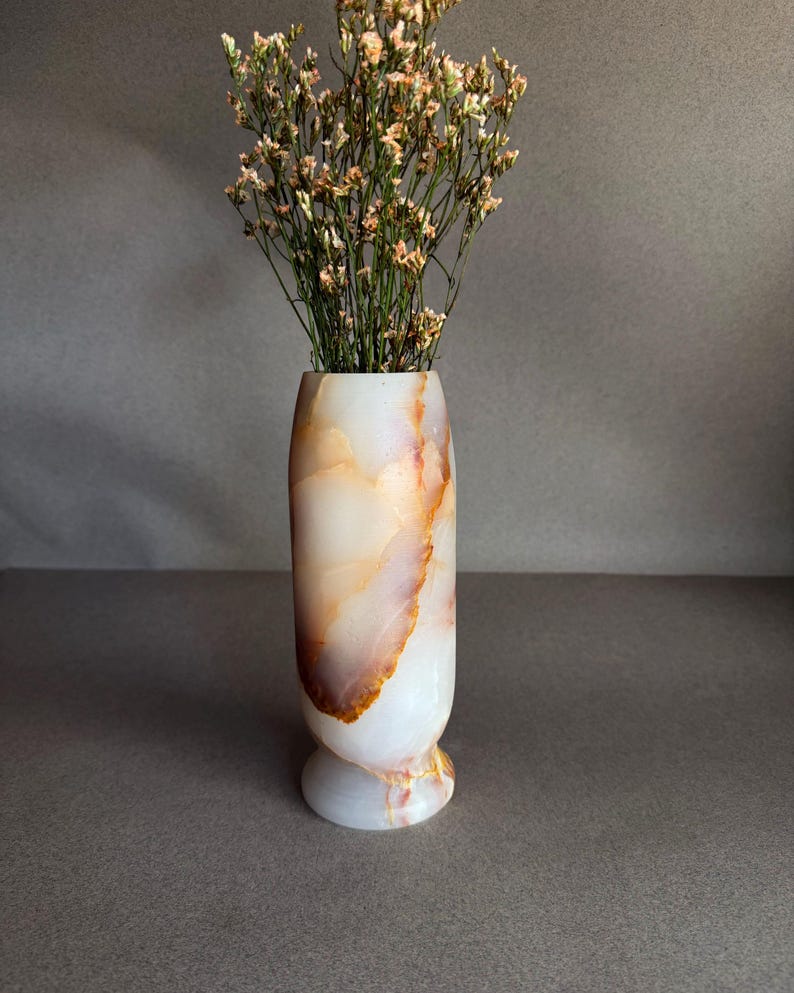 Pode incluir: Um vaso decorativo de &ocirc;nix branco e laranja com a parte superior alargada e uma base arredondada. O vaso &eacute; preenchido com flores secas com pequenas flores de cor clara. O vaso &eacute; um elemento decorativo para casa.