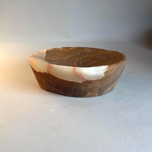 May include: A handcrafted wooden bowl with a unique design. The bowl features a dark brown exterior with a lighter, cream-colored band around the middle. The interior is a smooth, dark brown. The bowl is oval-shaped and sits on a neutral surface.