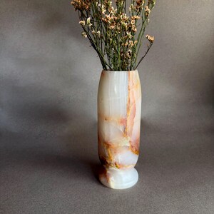 Pode incluir: Um vaso decorativo feito de &oacute;nix branco e com veios laranja. Cont&eacute;m um buqu&ecirc; de flores secas em tons de bege e castanho. O vaso tem uma base arredondada e um acabamento polido, ideal para decora&ccedil;&atilde;o de interiores.