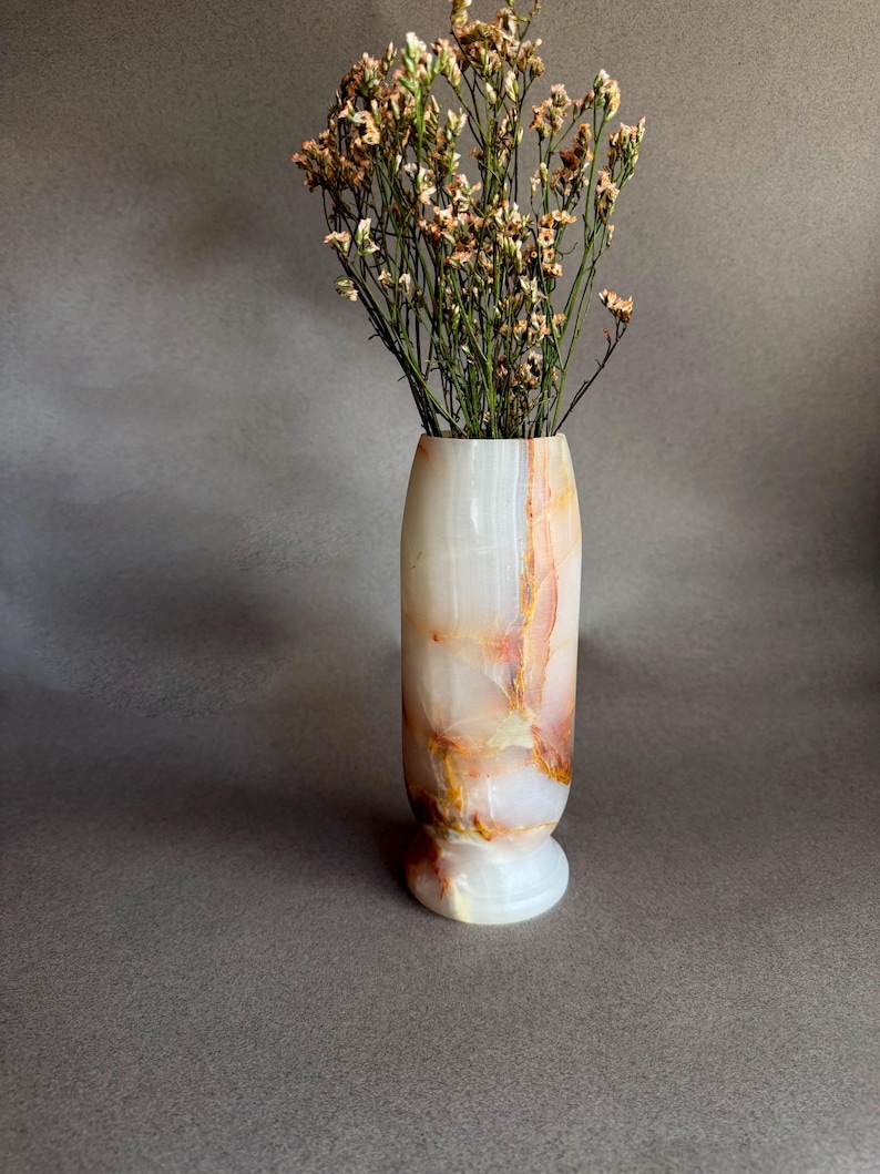 Pode incluir: Um vaso decorativo feito de &oacute;nix branco e com veios laranja. Cont&eacute;m um buqu&ecirc; de flores secas em tons de bege e castanho. O vaso tem uma base arredondada e um acabamento polido, ideal para decora&ccedil;&atilde;o de interiores.