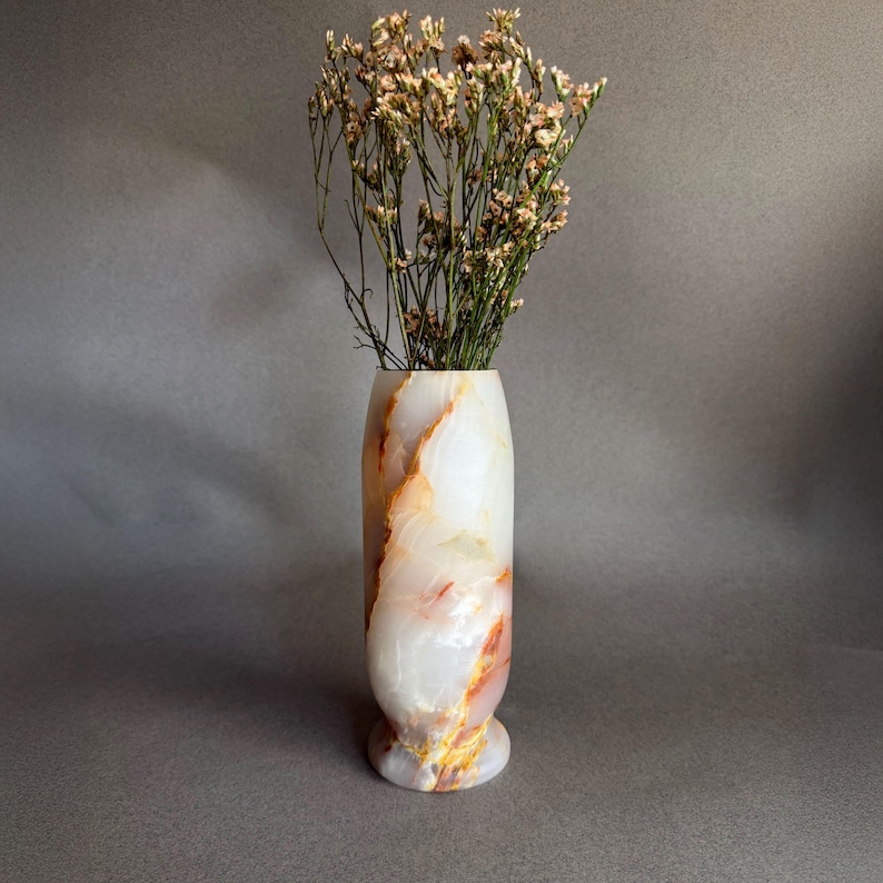 Pode incluir: Um vaso de &ocirc;nix branco e laranja com flores secas. O vaso tem uma base arredondada e um corpo cil&iacute;ndrico com veios laranja e dourados. As flores secas s&atilde;o de cor bege claro e adicionam um toque natural.