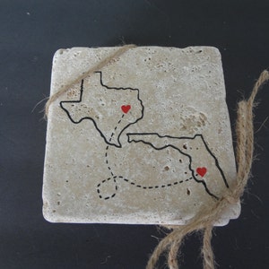 May include: A beige stone coaster with a black outline of Texas and Florida with a dotted line connecting the two states. There are two red hearts on the states.