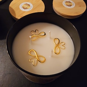 Könnte beinhalten: Eine weiße Kerze mit drei Dochten und drei goldenen Unendlichkeits-Herz-Anhängern oben. Die Kerze befindet sich in einem schwarzen Glas mit einem weißen Etikett.