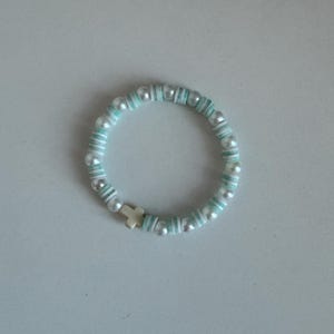 May include: A beaded bracelet featuring alternating white faux pearls and light blue disc beads. A small gold-colored cross charm is centered on the bracelet. The bracelet is designed for wrist wear.