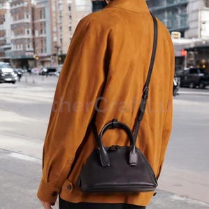 May include: A black leather handbag with a top handle and a long strap. The bag is worn by a person wearing a brown suede jacket.