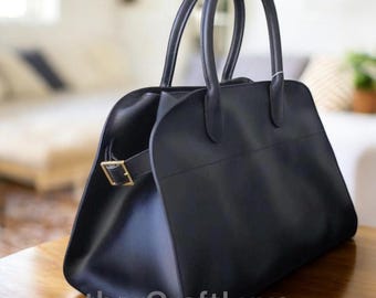 May include: A black leather handbag with two rounded handles and a gold-colored buckle closure. The bag has a structured shape and a minimalist design, suitable for everyday use. The bag is sitting on a wooden surface.