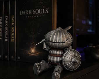 May include: A silver-toned, onion-shaped knight figurine in articulated armor, holding a small sword and a round shield. Behind the figure stands a Dark Souls Trilogy game case with the text Dark Souls, Dark Souls II, and Dark Souls III visible.