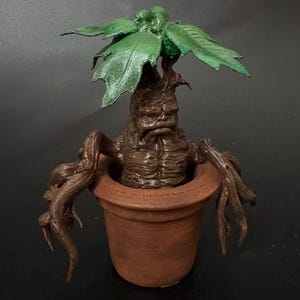 May include: A detailed, brown Mandrake plant figurine in a terracotta pot. The plant has a humanoid form with a distressed face, long roots, and large green leaves. The pot is round and unglazed, set against a dark background.