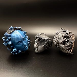 Peut inclure: Trois sculptures abstraites de visages humains. L'une est bleue avec des formations cristallines, une autre est grise avec un motif en spirale et la troisième est grise avec un aspect vieilli. Les sculptures sont posées sur un fond sombre.