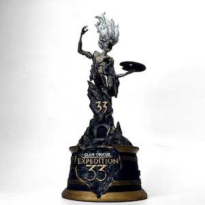 May include: A dark sculpture of a figure with flowing hair, holding a palette. The sculpture is mounted on a base with the text "Clair Obscur Expedition 33" in gold lettering. The figure is standing on a rocky structure.