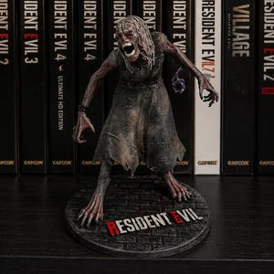 May include: A detailed figurine of a creature with pale, textured skin and long, stringy hair, wearing a tattered, dark gray dress. The figure stands on a circular base with the text Resident Evil, positioned in front of a row of video game cases.